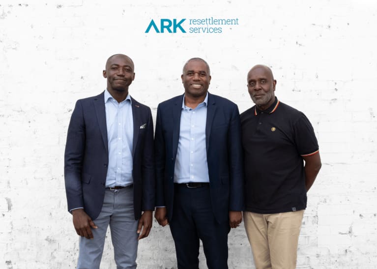 Three men standing together against a weathered white wall with ARK resettlement services logo