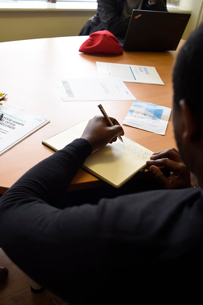 Person writing in notepad at table with laptop, documents, and red hat visible