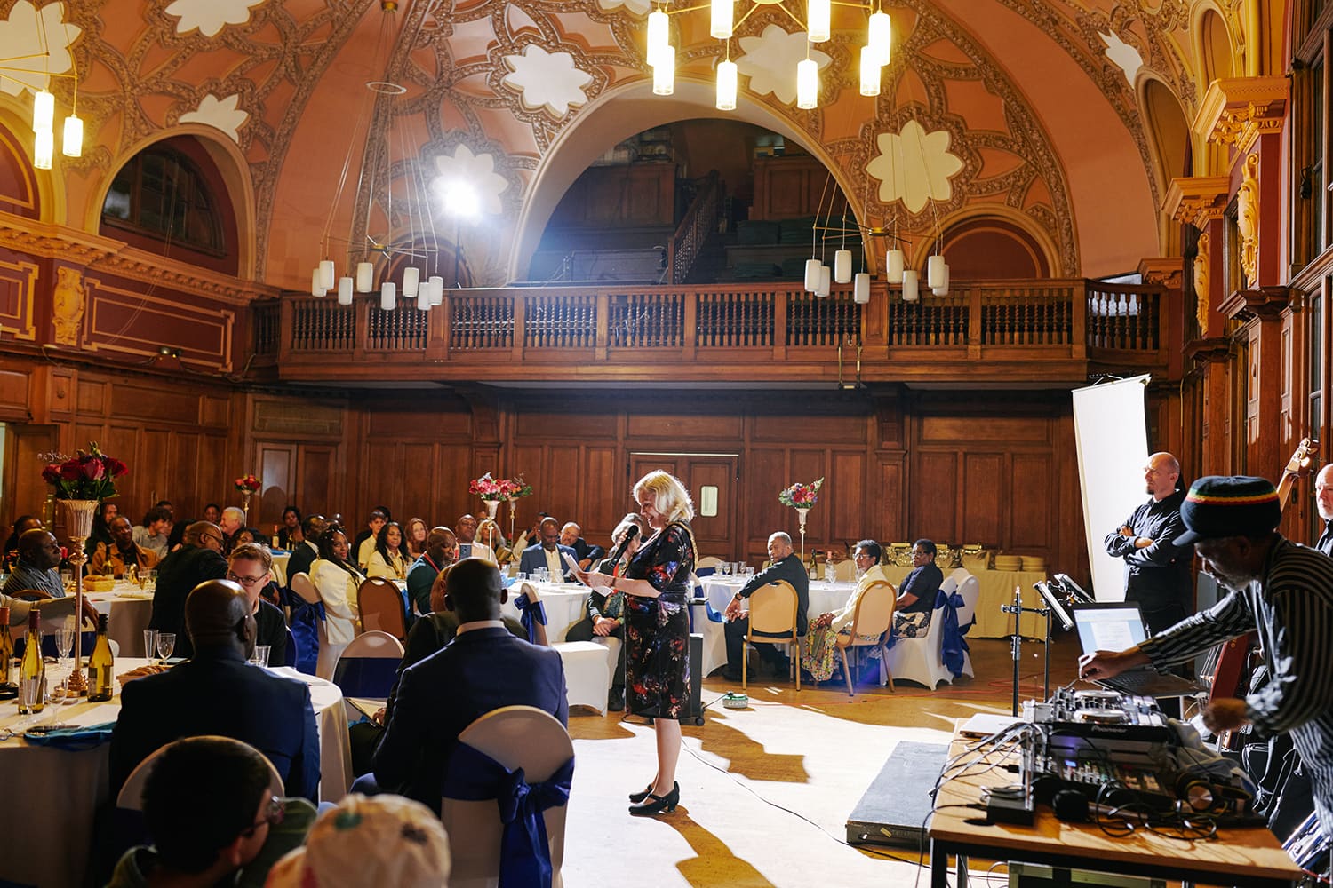 Woman speaking at podium in ornate wooden hall with seated guests at tables and band members