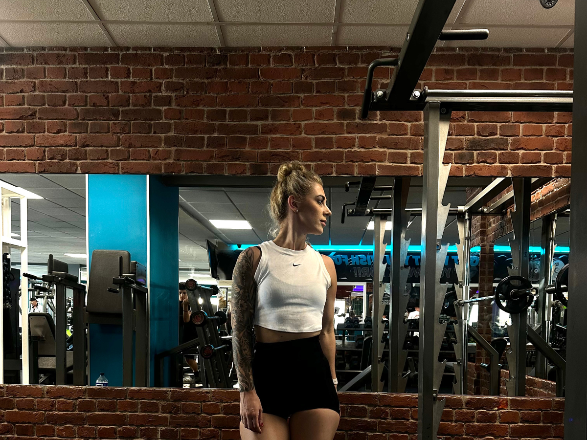 Woman in white crop top and black shorts standing in gym with brick walls and blue neon lighting