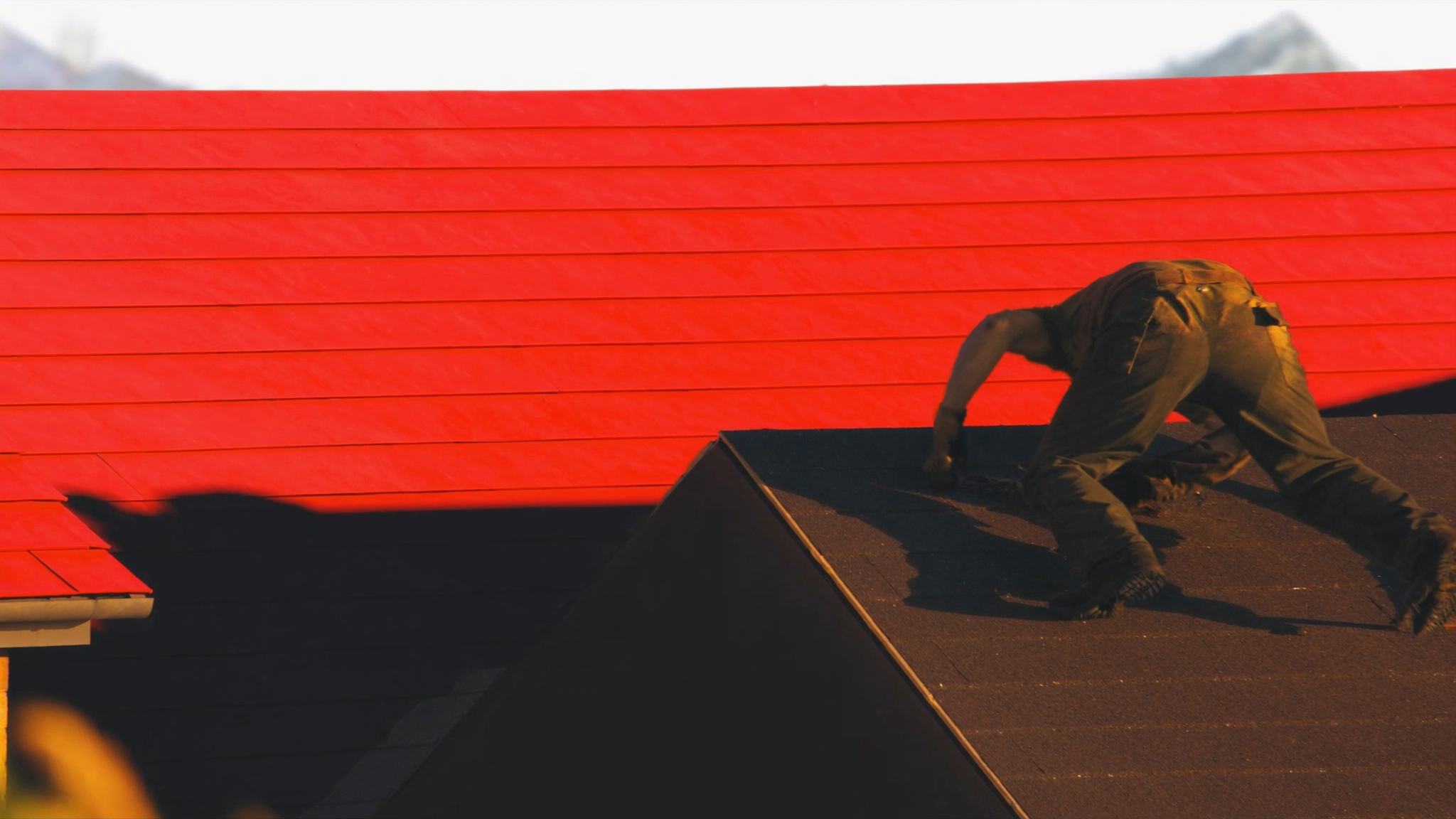 Worker in brown clothing crouching on dark roof beside bright red metal roofing panels