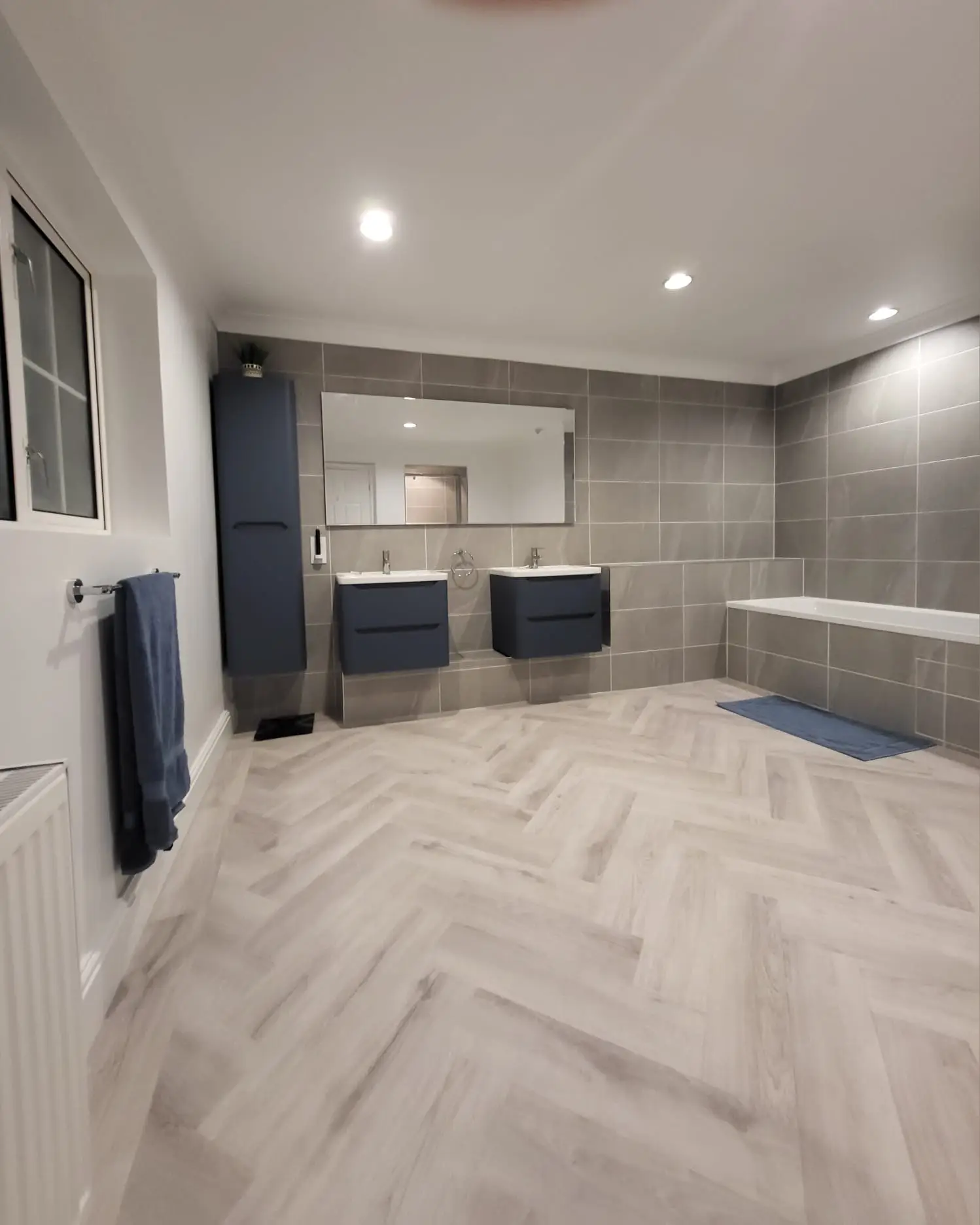 Modern bathroom with dual dark vanities, large mirror, gray tile walls, and herringbone wood-look flooring