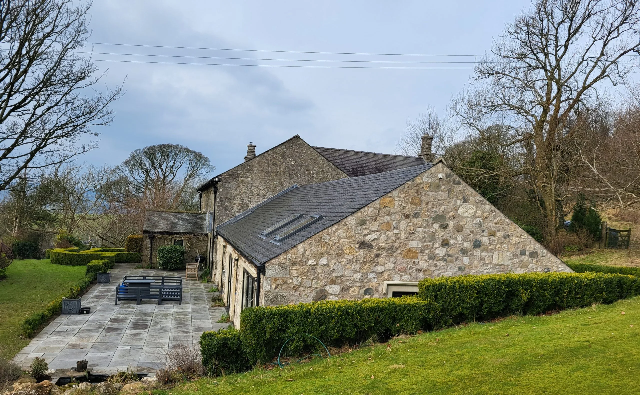 Modern stone cottage with slate roof, patio area, and surrounding hedges and grass