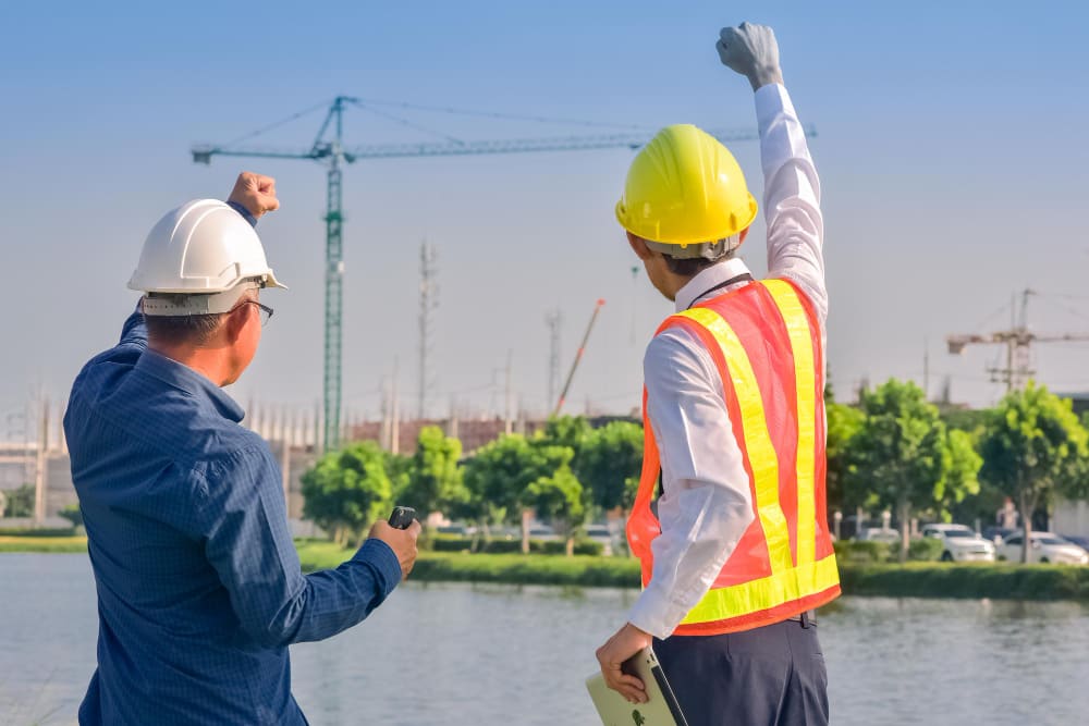Two construction workers in safety gear observing large industrial site with cranes and water