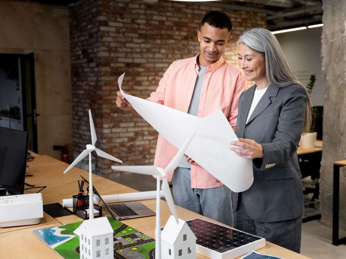 Two professionals reviewing blueprint in office with renewable energy models on desk