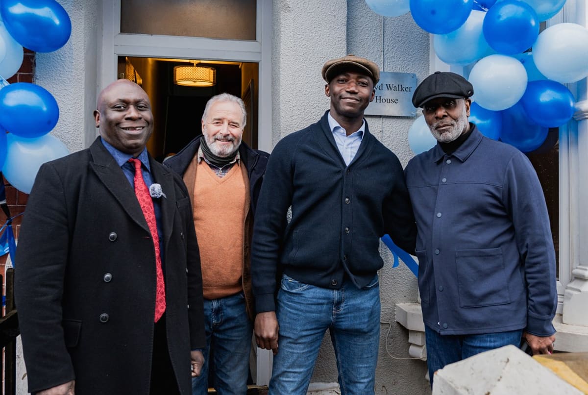 Four men standing together outside a building entrance with blue and white balloons