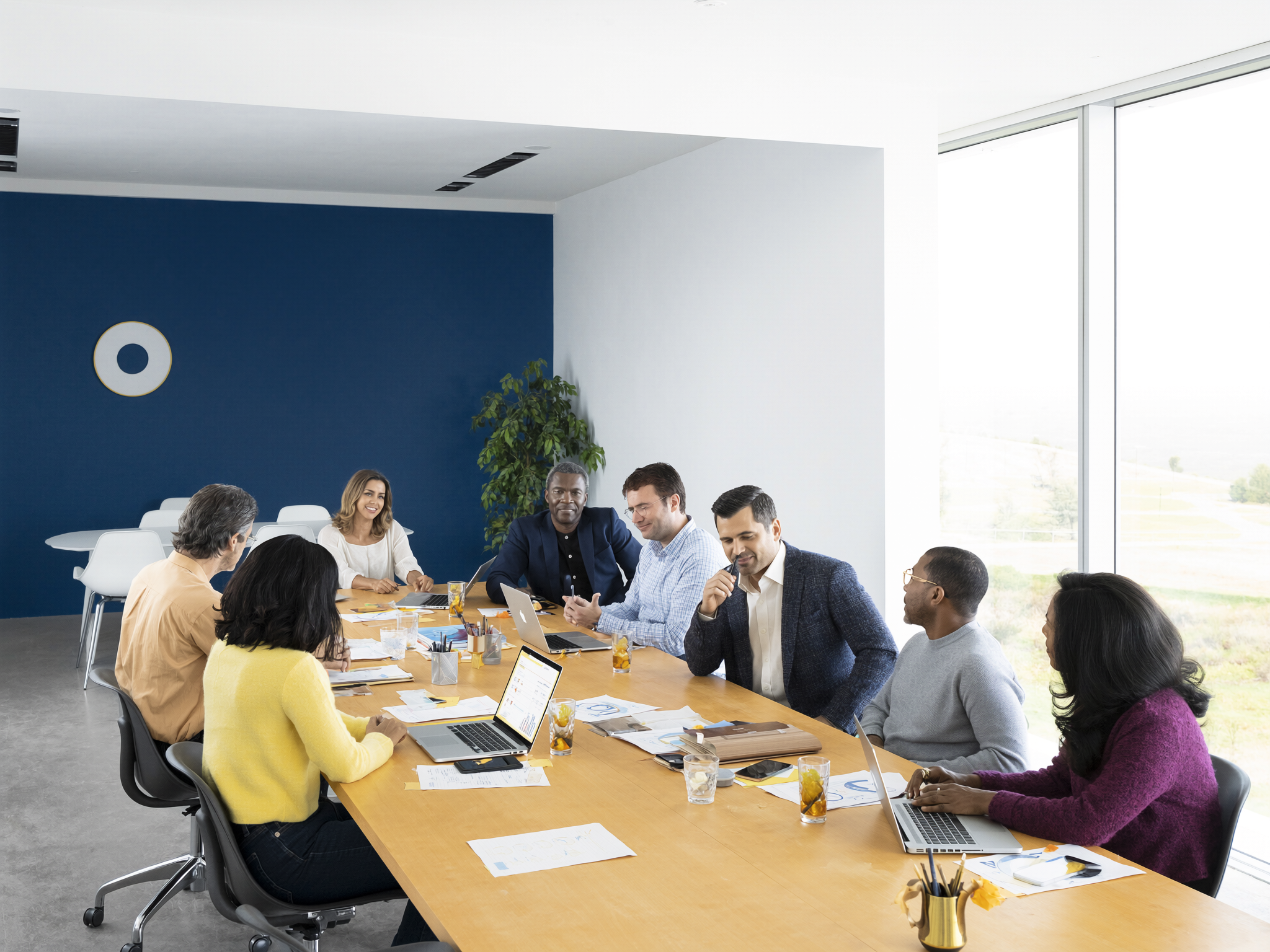 Diverse team of seven people meeting around wooden table in modern office with deep blue wall