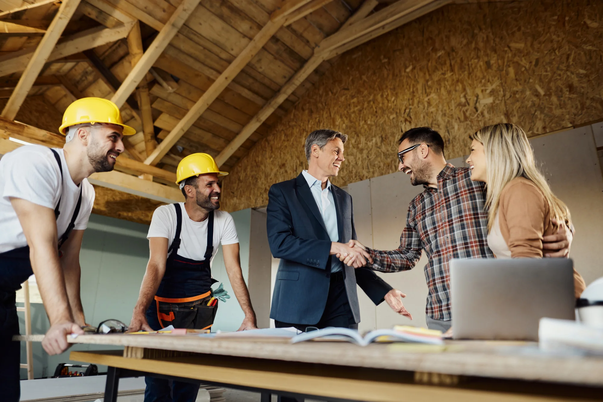Construction workers and business people shaking hands at table with blueprints indoors