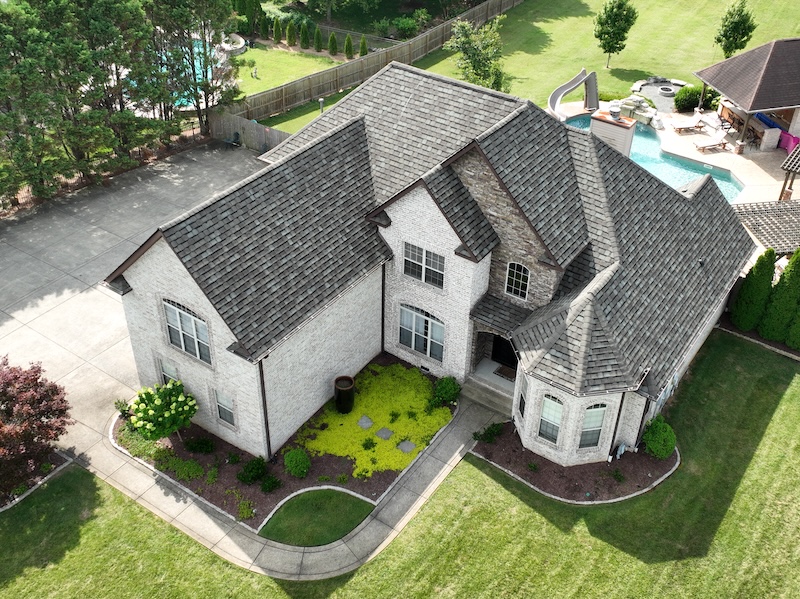 Aerial view of two-story white home with gray roof shingles, driveway, and landscaping