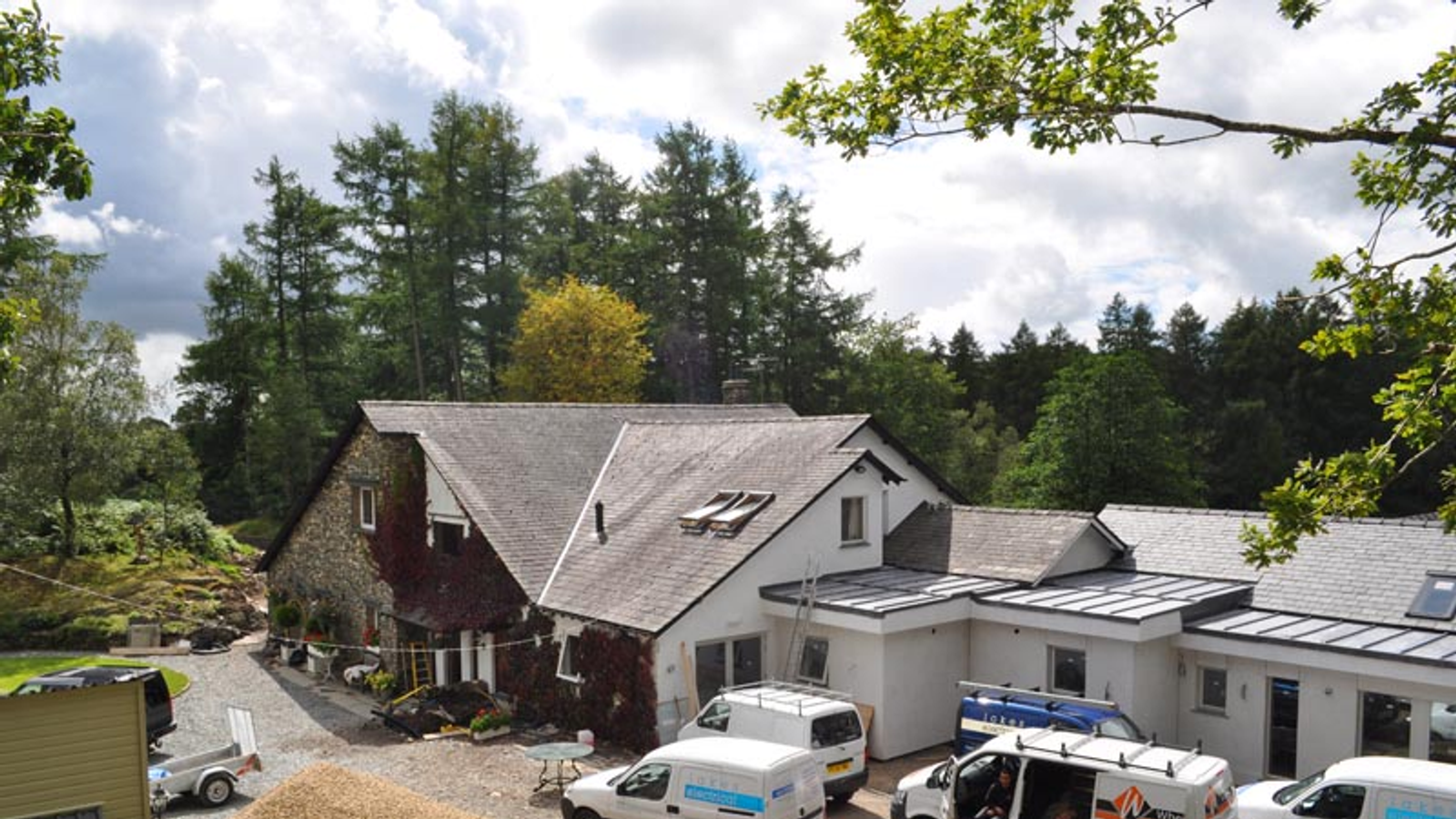 Aerial view of residential property with stone and white buildings, parked vehicles, and surrounding forest