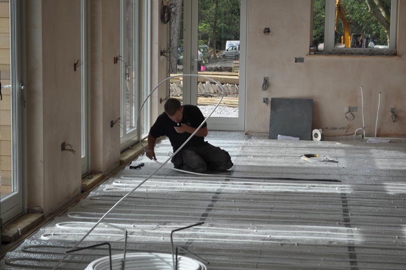 Worker installing underfloor heating pipes in room under construction with large windows.