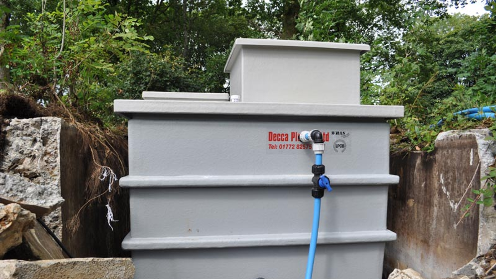 Gray plastic water tank with blue pipe valve, surrounded by trees and concrete blocks