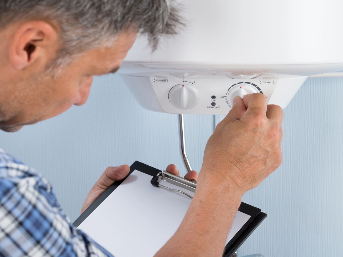 Man adjusting temperature dial on wall-mounted boiler unit while holding clipboard