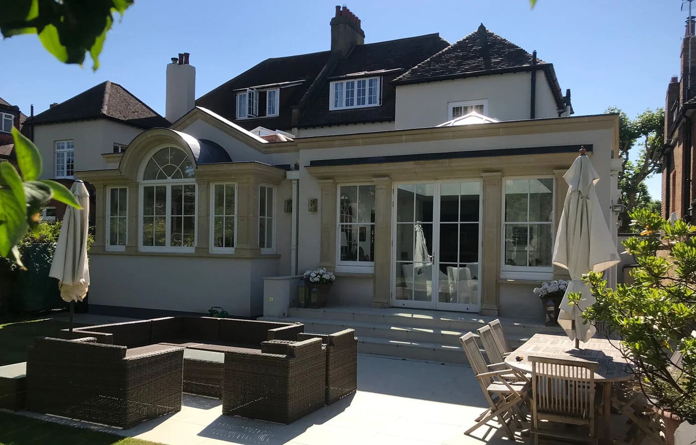 Cream-painted residential extension with large windows, patio furniture, and mature house behind