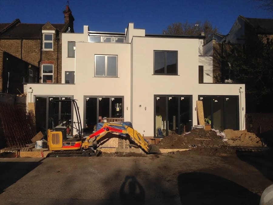 Modern cream residential extension under construction with black windows and excavator in foreground